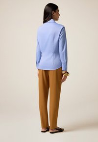 Donna di spalle che indossa una camicia a maniche lunghe azzurro chiaro, pantaloni marroni, scarpe nere slip-on e un grande braccialetto d'oro al polso destro.
