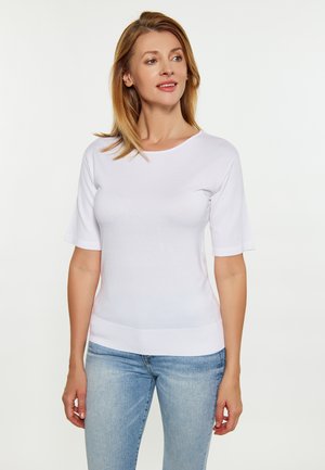 Kvinde med lysbrunt hår iført en simpel hvid t-shirt med korte ærmer og lyseblå jeans, der kigger lidt til siden.