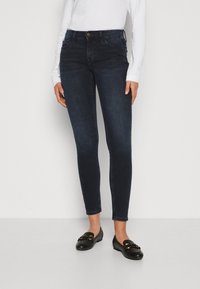 Jeans ajustados de un azul oscuro, confeccionados en denim, con una cintura de altura media, diseño de cinco bolsillos y cierre de botón. Se combinan con mocasines negros.