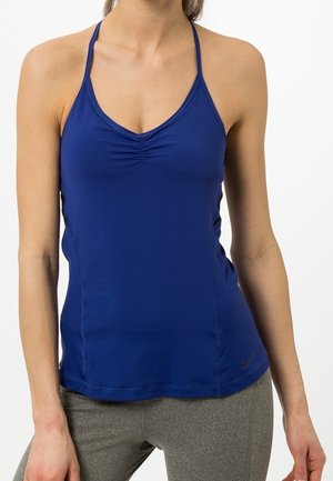 Frau trägt ein blaues ärmelloses Sporttop mit gerafftem Vorderteil und graue Leggings vor weißem Hintergrund.