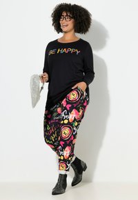 Glunderende vrouw met krullend haar draagt een zwart shirt met de tekst "BE HAPPY", kleurrijke patroonbroek, gestifte zwarte laarzen en houdt een zilveren pailletten tas vast.