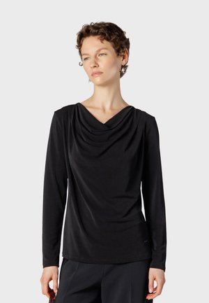 Femme aux cheveux courts et bouclés portant un haut noir à manches longues avec un décolleté drapé sur un fond clair uni.