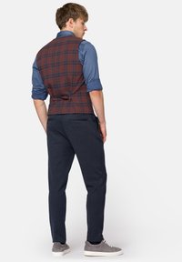 Brązowa kamizelka w kratę o dopasowanym kroju, noszona na niebieskiej koszuli. Ciemnogranatowe spodnie i szare sneakersy dopełniają strój.