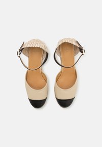 Scarpe a zeppa in tela beige con punta in pelle nera, cinturino in tessuto con motivo e fibbia alla caviglia. Texture liscia e forma arrotondata.