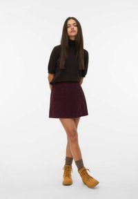 Jonge vrouw met lang bruin haar in een zwarte coltrui, bordeauxrode rok, grijze sokken en beige veterschoenen, staande tegen een witte achtergrond.