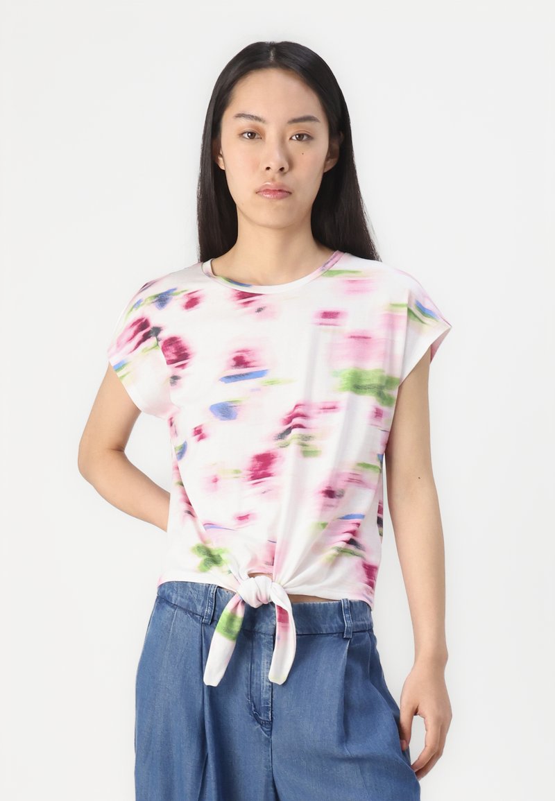 Kurze, weiße T-Shirt mit einem Knoten in der Taille, das ein farbenfrohes abstraktes Muster in Pink, Grün und Blau aufweist. Glatte Stofftextur.