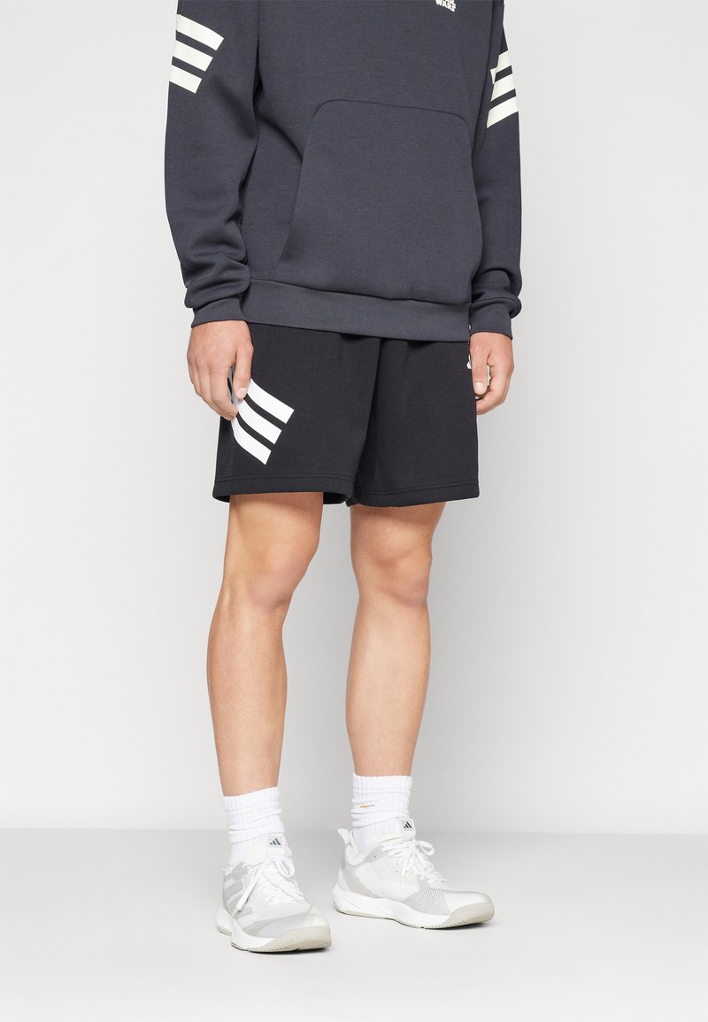 Mørk grå hettegenser med hvit logo, svarte shorts med hvite striper, og grå treningssko. Hvite ankelsokker fullfører looken.