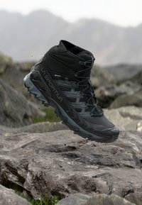 Bota de caminhada de cano alto preta com parte superior sintética texturizada, forro Gore-Tex e sola multidirecional, mostrada no ar acima de um terreno rochoso.