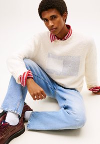 Κρεμ πλεκτό πουλόβερ με μπλε μπαλώματα, κόκκινο-λευκό ριγέ πουκάμισο από κάτω, ανοιχτό μπλε τζιν και μπορντό δερμάτινα παπούτσια με κορδόνια.