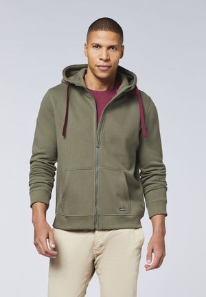 Olivgrüner Zip-Hoodie aus strukturiertem Stoff, mit Kordeln und seitlichen Taschen. Getragen über einem kastanienbraunen Shirt, kombiniert mit beigen Hosen.