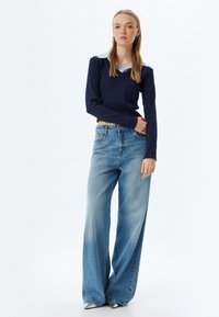 Marineblaue Langarmbluse mit V-Ausschnitt und einem hellblauen Hemd darunter, kombiniert mit hellblauen, weiten Jeans und silbernen Schuhen.