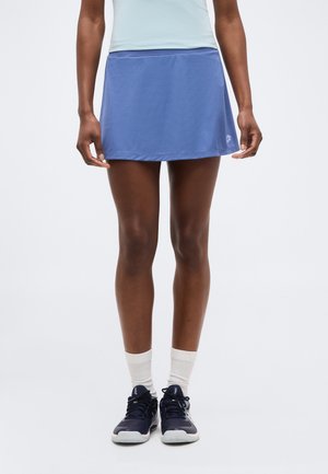 Άτομο που φοράει ανοιχτό μπλε αθλητική φούστα, λευκές κάλτσες και μαύρα αθλητικά παπούτσια, στέκεται μπροστά σε απλό λευκό φόντο.