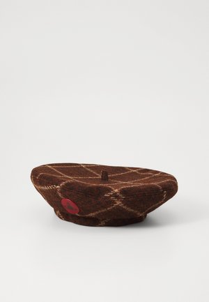 Béret en laine marron avec un motif à carreaux, surmonté d'un petit bouton central et d'un patch logo rouge sur le côté. Texture douce, forme plate et ronde.