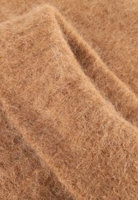 Weiche, flauschige Stoff in Kamelbraun mit einer strukturierten, wolligen Oberfläche und sichtbaren Fasern, die in sanften Falten angeordnet sind.