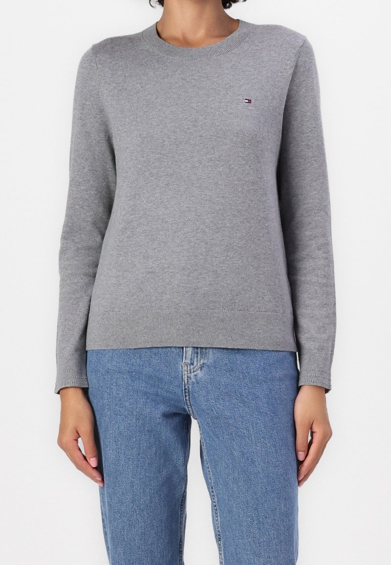 Femme portant un pull gris uni à manches longues avec un petit logo sur la poitrine et un jean en denim bleu clair, debout les bras détendus.