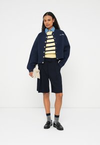 Námornícky skrátený kabát s zlatými gombíkmi, žltý pruhovaný sveter, strihané tmavé šortky, sivé ponožky, čierne mokasíny a béžová kabelka.