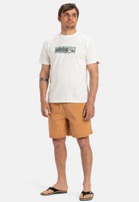 Camiseta de algodón blanca con logo verde, combinada con pantalones cortos naranjas. La persona lleva sandalias negras y muestra un tatuaje en el brazo.