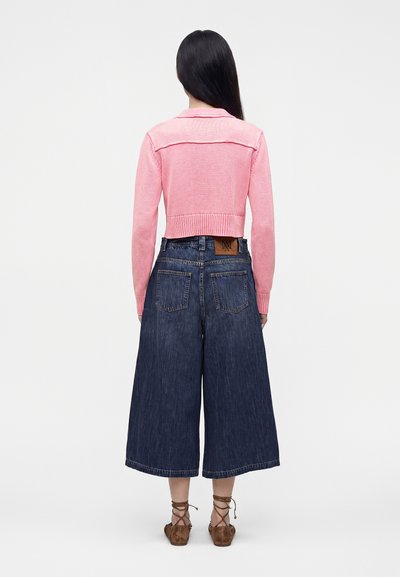 Femme aux longs cheveux foncés, portant un pull court rose, un pantalon large en denim bleu foncé et des chaussures plates marron à lacets, debout, de dos.