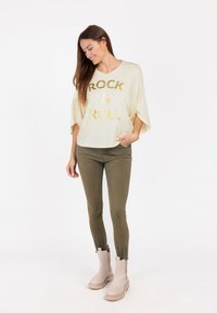 Top a V con scollo a V crema con la scritta "ROCK & ROLL" in lettere dorate, abbinato a pantaloni aderenti verde oliva e stivaletti beige alla caviglia.