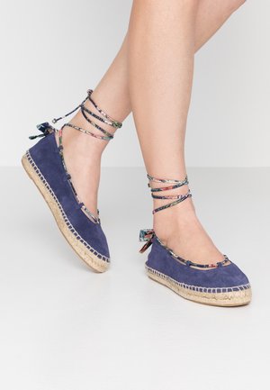 Benen met blauw suède espadrilles met bloemenprint enkelwikkellinten en jute plateauzolen, staand op een lichtgrijze vloer.