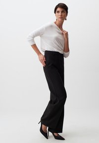 Vrouw in een witte longsleeve top, zwarte pinstripe wijde broek en zwarte hoge hakken, poserend tegen een effen witte achtergrond.