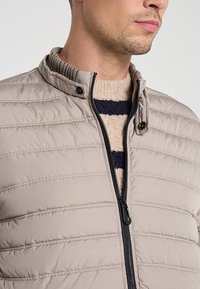 Lichtbeige gewatteerde donsjas met ritssluiting aan de voorkant en een geribde kraag, gedragen over een crème- en marineblauw gestreepte trui.