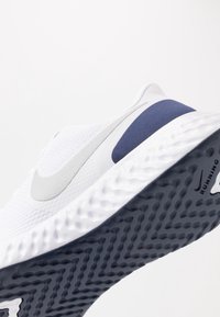 Zapatilla de correr blanca y azul marino con suela texturada, logo de Nike en gris claro y parte superior de malla, mostrada en ángulo sobre un fondo sencillo.