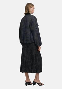 Black bomber jacket with textured fabric, ribbed collar, and utility pocket. Paired with a black pleated midi skirt and heeled sandals.