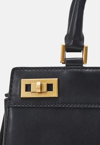 Zwarte leren handtas met rechthoekige goudkleurige draaivergrendeling en afgewerkt handvat met stiksels op witte achtergrond.