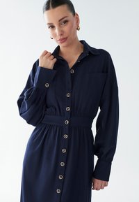 Abito utility blu navy con maniche lunghe a sbuffo, davanti con bottoni, cintura in vita e dettagli con bottoni color oro. Tessuto liscio con vestibilità comoda.