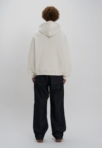 Sudadera de gran tamaño color crema con capucha ajustable, combinada con pantalones de mezclilla oscuros de pierna ancha y zapatos marrones. Diseño minimalista y textura suave.