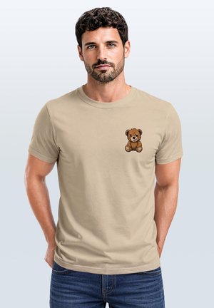 Man with short curly hair and beard wearing beige t-shirt with small embroidered brown teddy bear on chest, standing with hands in pockets.
