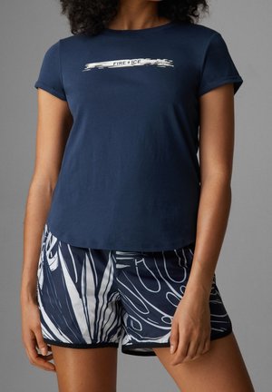 Kobieta w granatowym T-shircie z napisem "FIRE + ICE" oraz granatowych szortach z białymi abstrakcyjnymi wzorami liści.