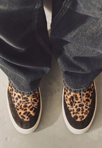 Chaussures à enfiler imprimé léopard en tissu texturé et daim, dotées d'une semelle en caoutchouc blanche, associées à un jean en denim foncé.