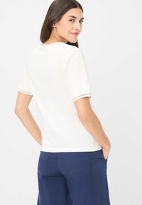 Witte top met korte mouwen en kantdetails op de mouwen, gecombineerd met blauwe broek met achterzakken en subtiele stiksels.