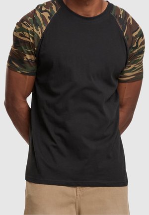 Mann trägt ein schwarzes T-Shirt mit camouflagegemusterten kurzen Ärmeln und hellbraunen Hosen, steht mit den Händen hinter dem Rücken.