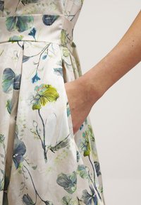 Kukkamekko kevyestä kankaasta, jossa on vihreitä ja sinisiä kasviprinttejä. Sivutasku näkyy, ja käsi ulottuu siihen, mikä korostaa toimintavuutta.