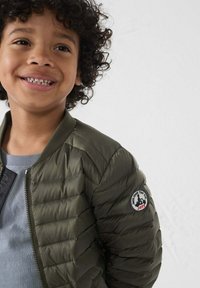 Giacca bomber imbottita verde con zip frontale, colletto a costine e patch del logo sulla manica. Indossata sopra una maglietta grigia chiara, con un look testurizzato.