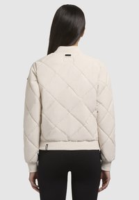 Béžová prešívaná bomber bunda s rebrovaným lemom, so stojacím golierom a štítkom na zadnej strane, z pohľadu zozadu.