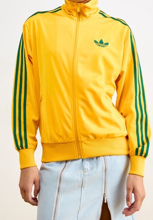 Personne portant une veste zippée jaune avec des rayures vertes sur les manches et un logo, associée à une jupe en jean bleu clair avec des fermetures éclair à l'avant.