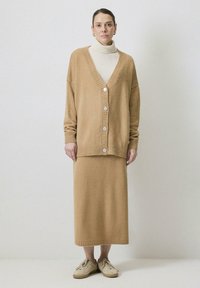 Tevebarna kötött kardigán bordás széllel és hat gombbal, egy hozzá illő, térd alá érő szoknyával párosítva. Egy világos garbóra viselve.