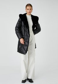 Vrouw in een zwarte pufferjas met een bontkraag, witte geribbelde trui, wijde witte broek en zwarte hakken tegen een effen achtergrond.