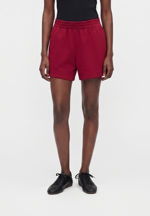 Person iført mørkerøde shorts med elastisk talje, sort ærmeløs top og sorte snørebåndssko, der står mod en ensfarvet baggrund.