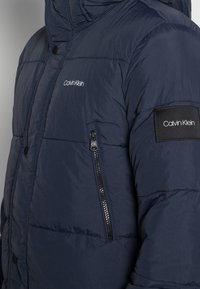 Granatowa kurtka puchowa z kieszenią na zamek, wysokim kołnierzem i naszywką z logo na rękawie. Widoczna gładka tekstura i pikowany design.