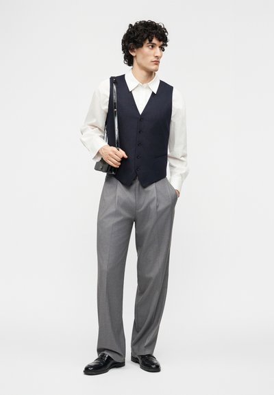 Άνδρας που φοράει μαύρο γιλέκο πάνω από λευκό πουκάμισο, γκρι παντελόνι, μαύρα παπούτσια, κρατώντας μαύρη τσάντα ώμου, στέκεται με το ένα χέρι στην τσέπη.