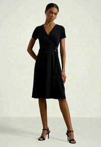 Γυναίκα που στέκεται φορώντας ένα κοντομάνικο μαύρο φόρεμα με ζώνη και μαύρα ψηλοτάκουνα σανδάλια σε απλή φωτεινή φόντο.