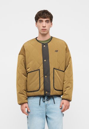 Junger Mann trägt eine hellbraune gesteppte Jacke mit schwarzem Besatz und großen Taschen über einem grünen Hemd und hellblauen Jeans, steht vor weißem Hintergrund.