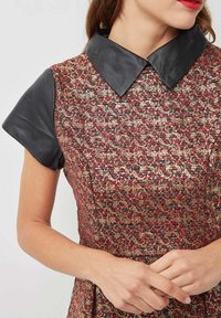 Vestido con un patrón texturizado en rojo y oro, con un cuello de cuero negro y mangas cortas, que combina elementos de diseño vintage y moderno.