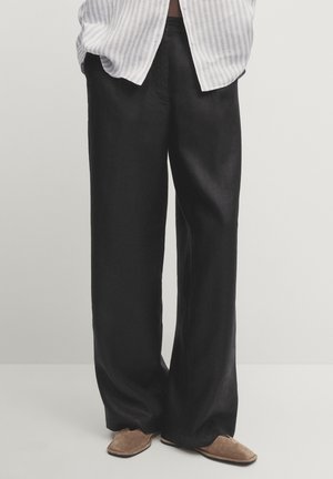 Personne portant un pantalon large noir ample, des chaussures plates marron et une chemise rayée blanche et grise partiellement visible.