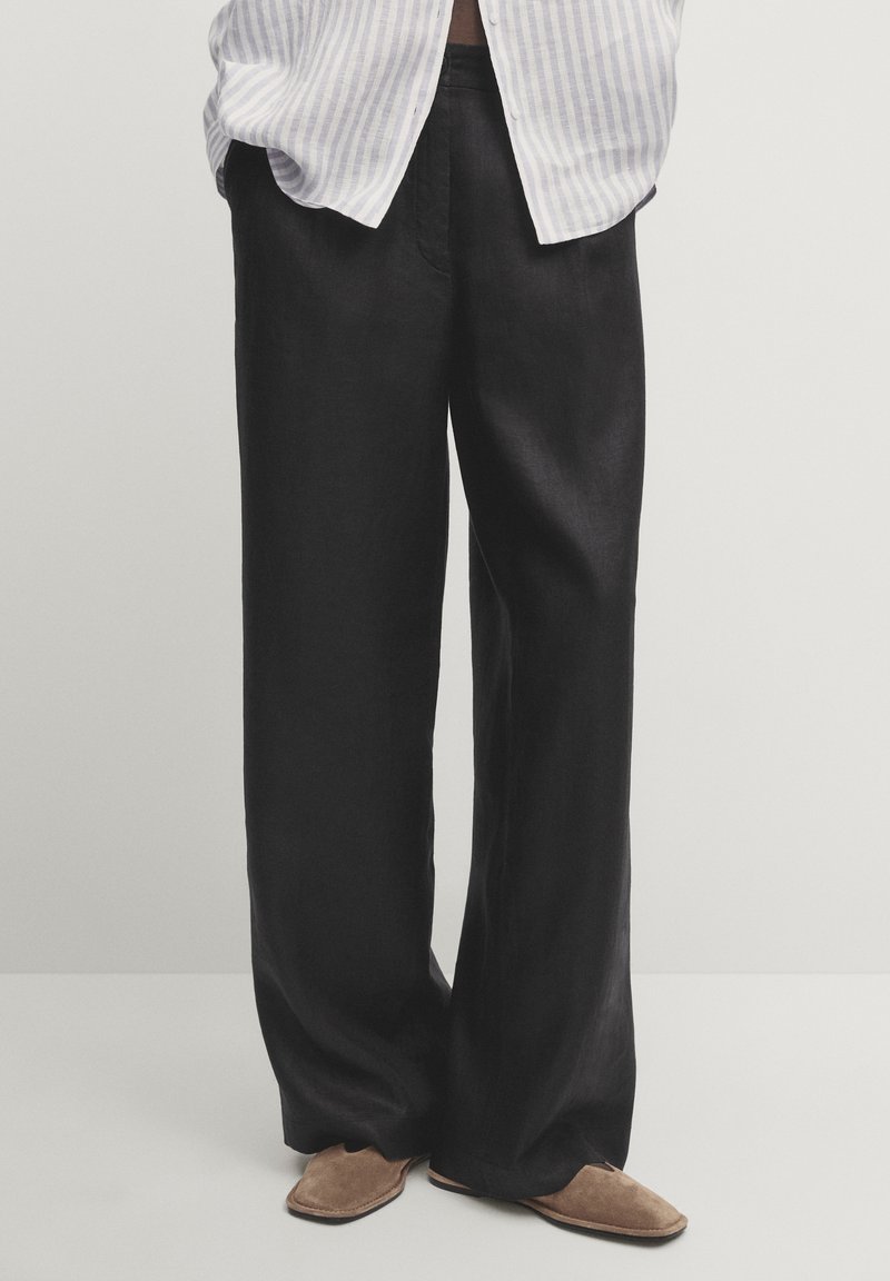 Personne portant un pantalon large noir ample, des chaussures plates marron et une chemise rayée blanche et grise partiellement visible.